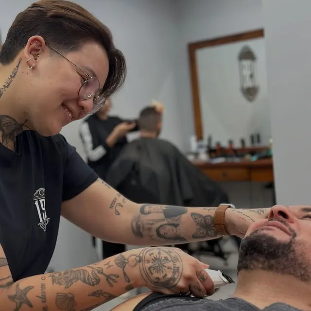 Barbeira de óculos e braços tatuados fazendo a barba de um cliente